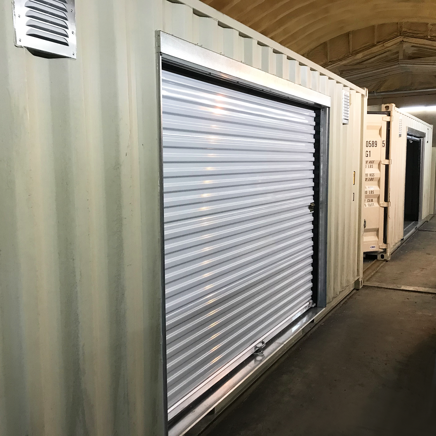 Side view of a roll-up door installed in a high cube shipping container using a galvanized steel framing kit. The door is securely fitted with a flush mount and clean trim, inside a covered storage facility. Framing kit only—door sold separately.