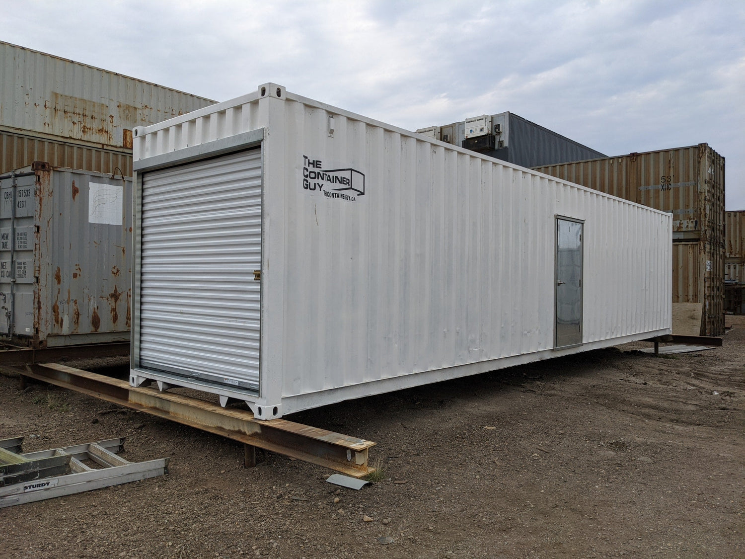 Modified white shipping container featuring a roll-up door on the end and a man door on the side, displayed in an outdoor container yard