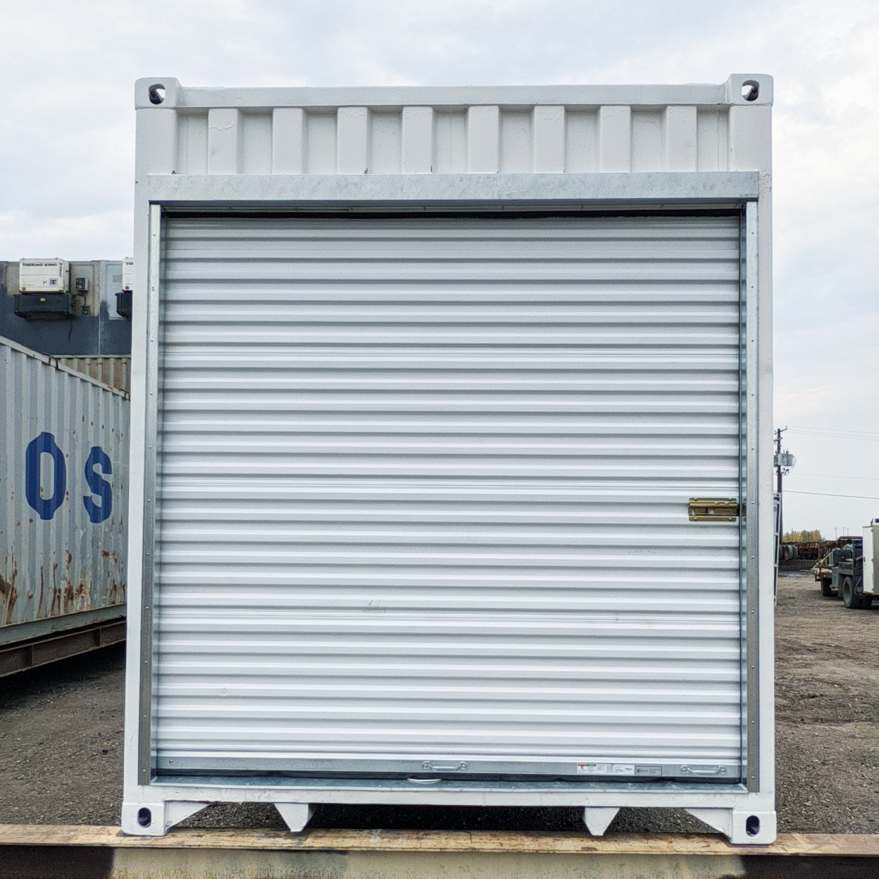 Roll-up door installed on the end of a white shipping container using a galvanized steel framing kit, photographed in an outdoor industrial yard.