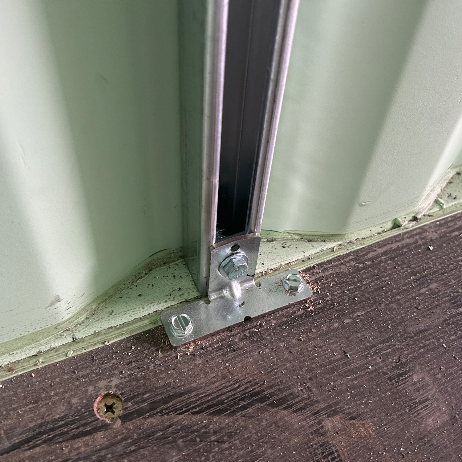 Close-up of a galvanized strut channel bracket mounted to the wooden floor and wall edge of a shipping container, securely anchoring a vertical strut for structural framing.