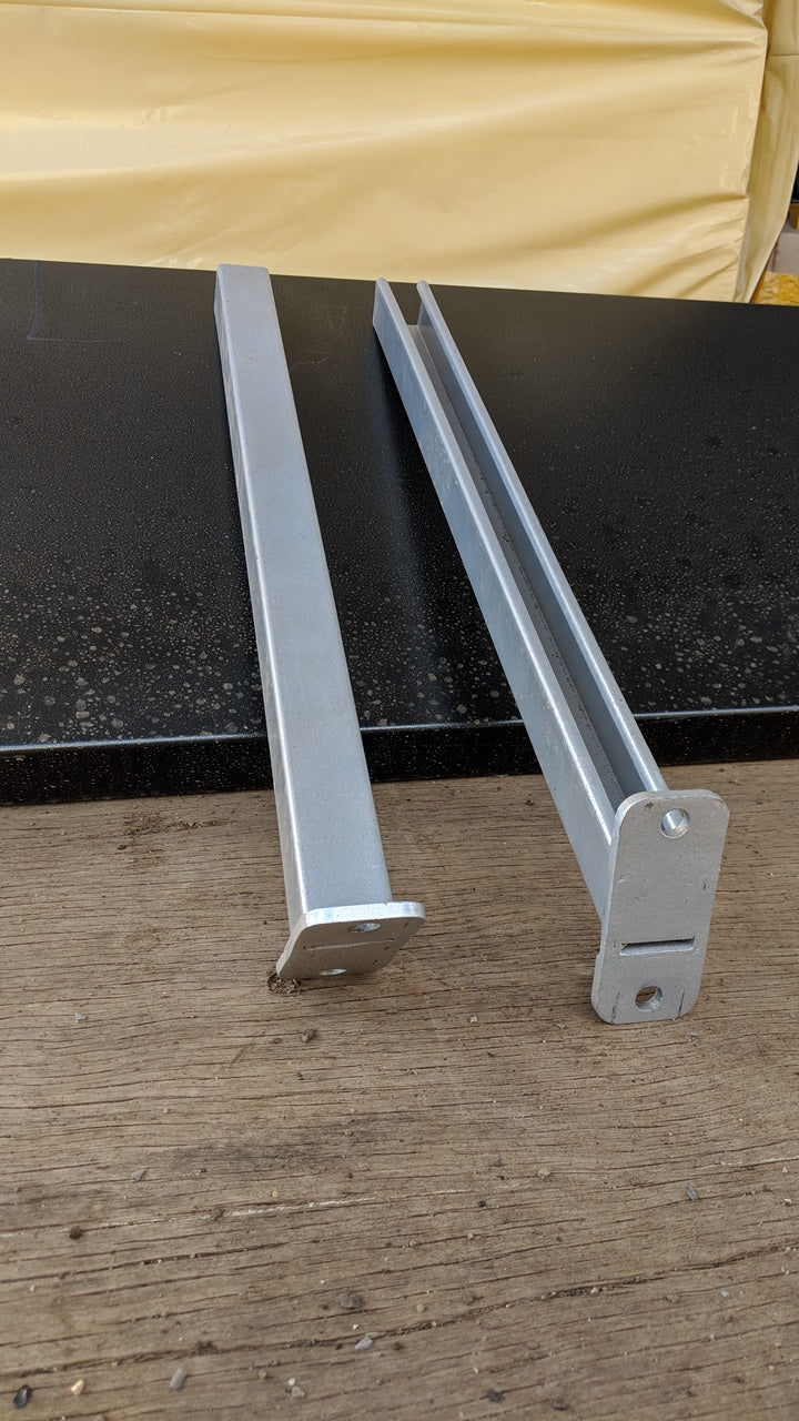 Two galvanized steel CSM Pipe Rack arms displayed outdoors on a wooden and metal surface, angled to show mounting plates and U-channel design for securing pipes inside shipping containers.