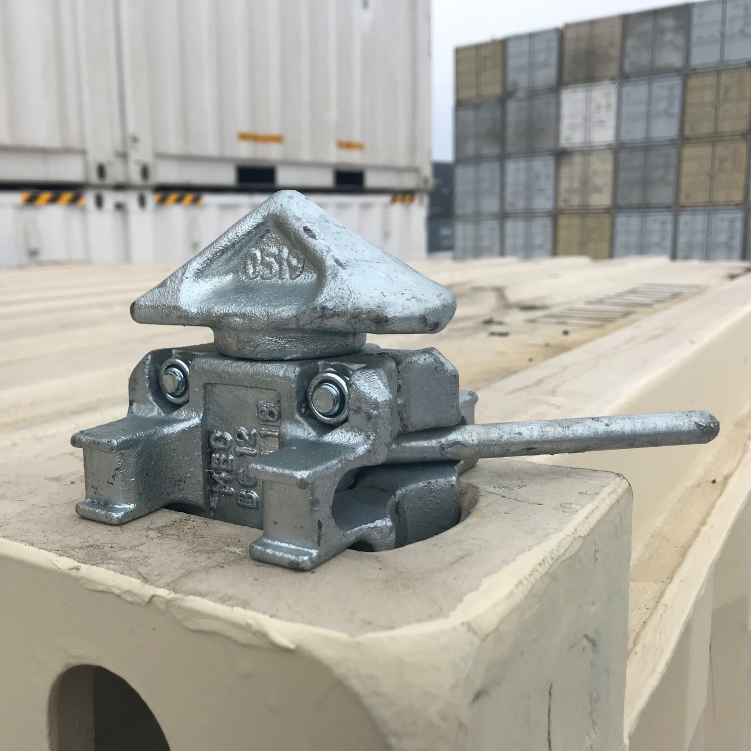 Shipping container twist lock installed on corner casting, securing stacked containers in a yard; heavy-duty galvanized steel construction with locking handle.